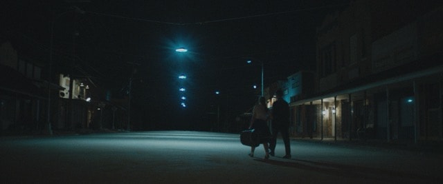 Two figures walking down a deserted small-town street at night under blue lights