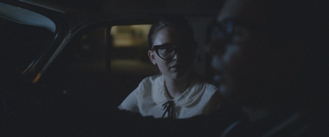 Young woman in cat-eye glasses looking tense in a dark car
