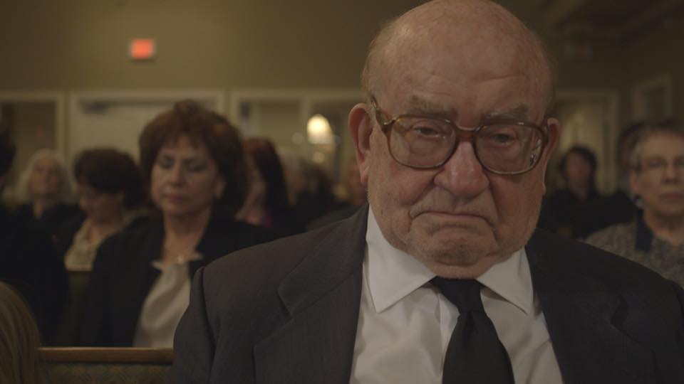 Ed Asner in suit and tie seated in church pew, deep in thought