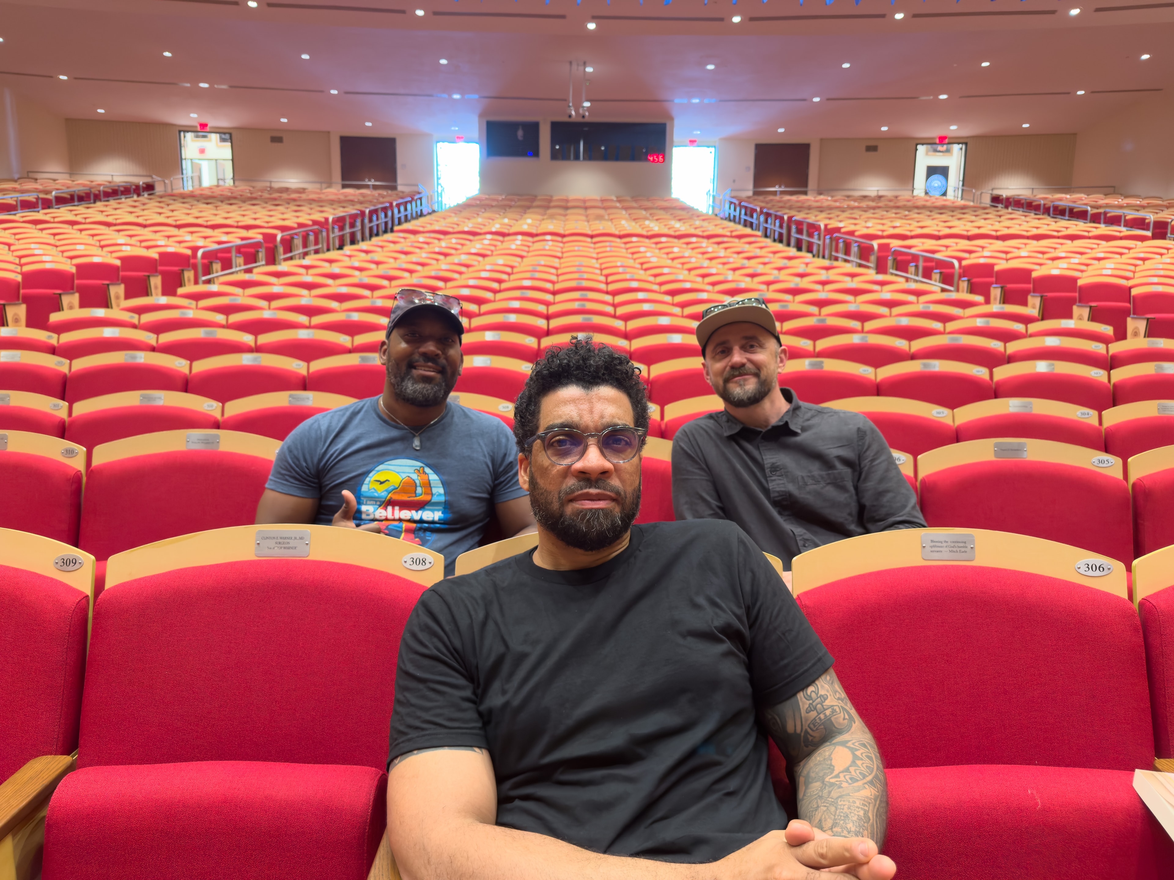 Three men seated in empty auditorium with red seats