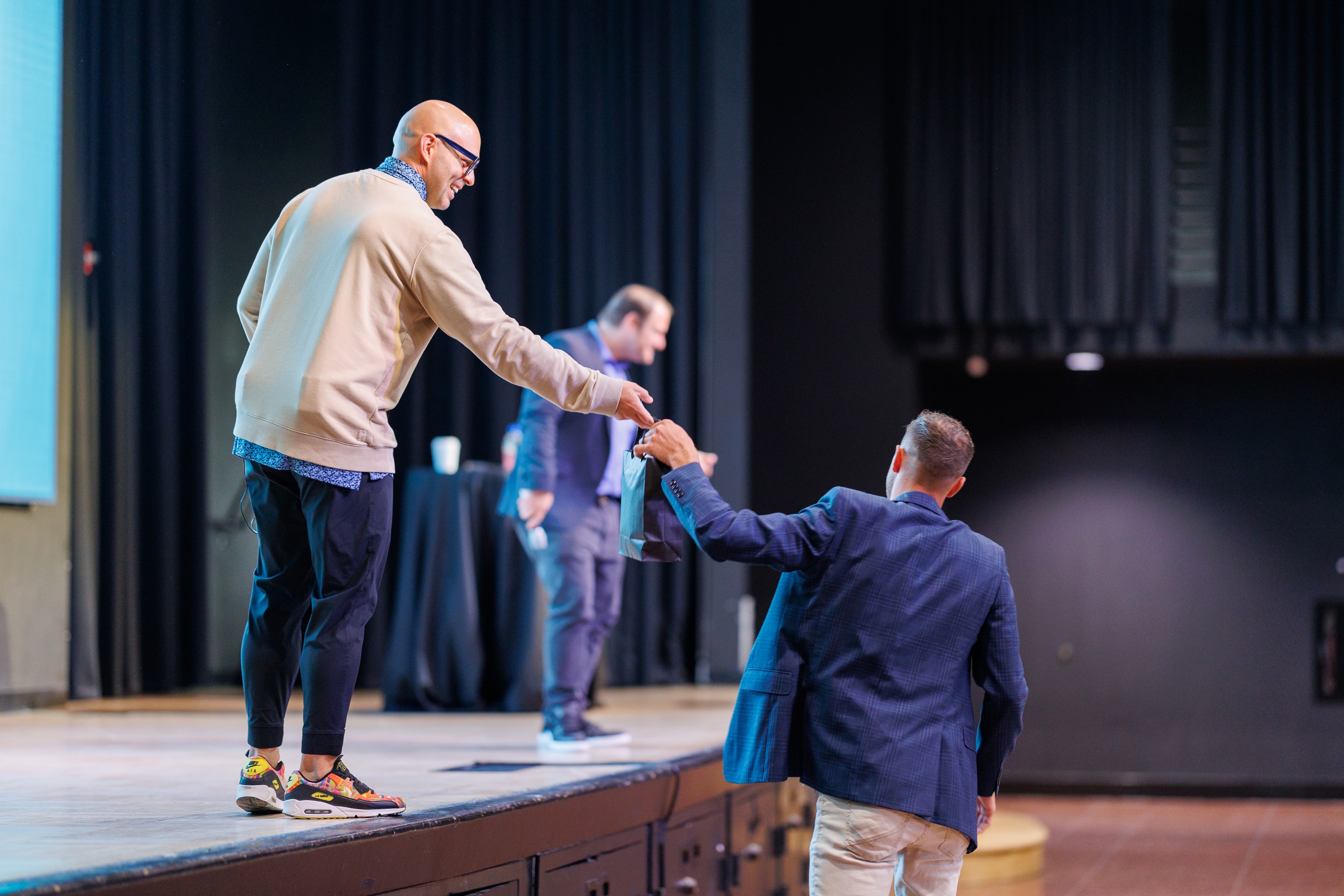Man on stage reaching down to hand something to person in audience