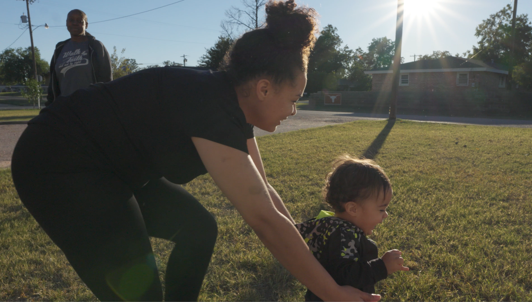 Mother helping toddler walk on grass in golden sunlight